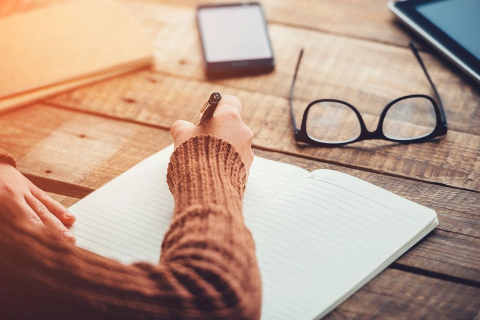 A person is writing on paper with glasses and phone